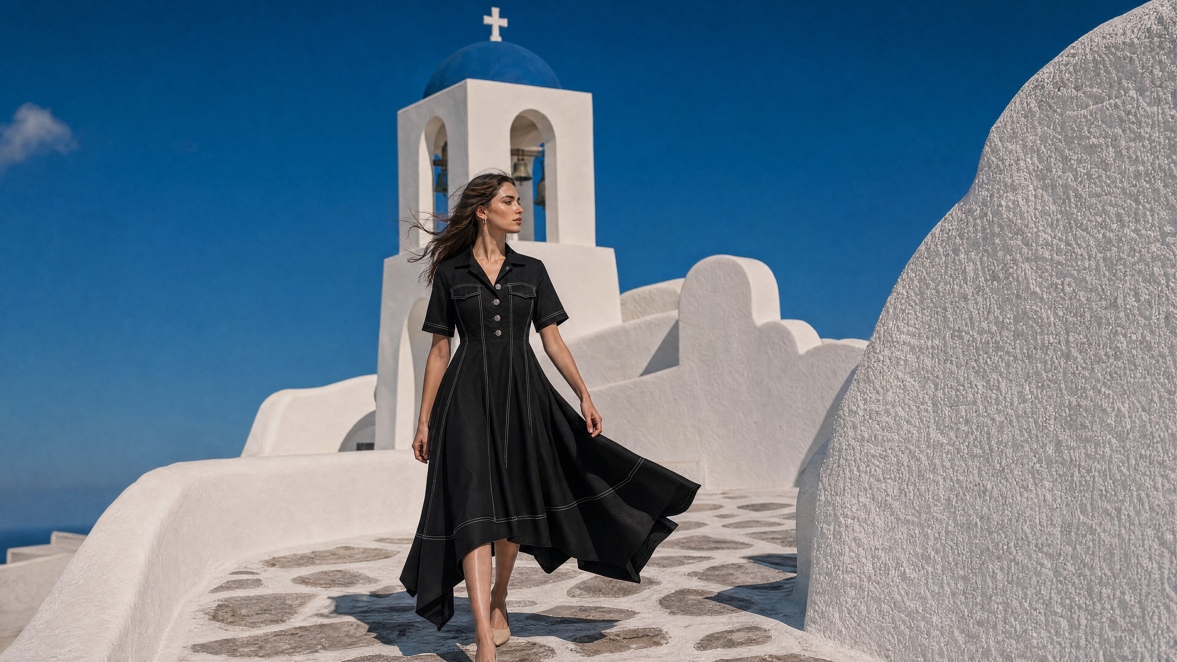 New Arrivals — model in a black pleated shirt dress walking a whitewashed path, blue-domed chapel behind